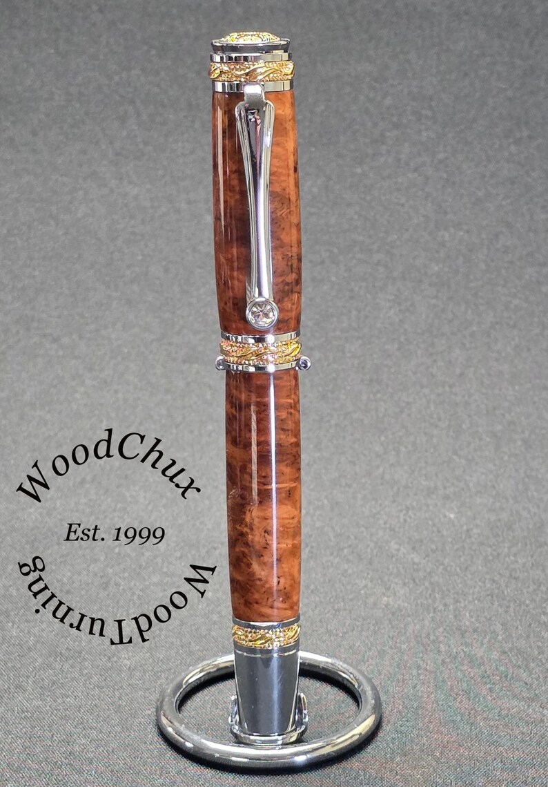 May include: A handcrafted pen with a polished, burl wood barrel, accented with gold and silver-tone metal. The pen features a silver clip and a decorative band with a clear gem. The pen is displayed on a silver stand. The text "WoodChux Est. 1999" is visible.