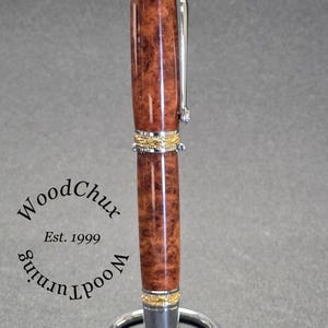May include: A handcrafted pen with a brown burl wood barrel and gold-colored accents. The pen features a silver clip and a decorative gold band. The pen is displayed on a silver pen stand. The text "WoodChux Est. 1999" is visible.