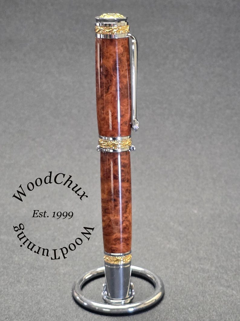 May include: A handcrafted pen with a brown burl wood barrel and gold-colored accents. The pen features a silver clip and a decorative gold band. The pen is displayed on a silver pen stand. The text "WoodChux Est. 1999" is visible.