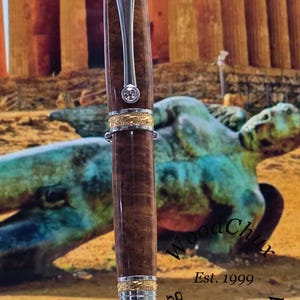 May include: A handcrafted wooden pen with a silver clip and gold accents. The pen features a dark brown, textured barrel and a sparkling gem. The pen is displayed against a blurred background with the text "Wood Turning Est. 1999".