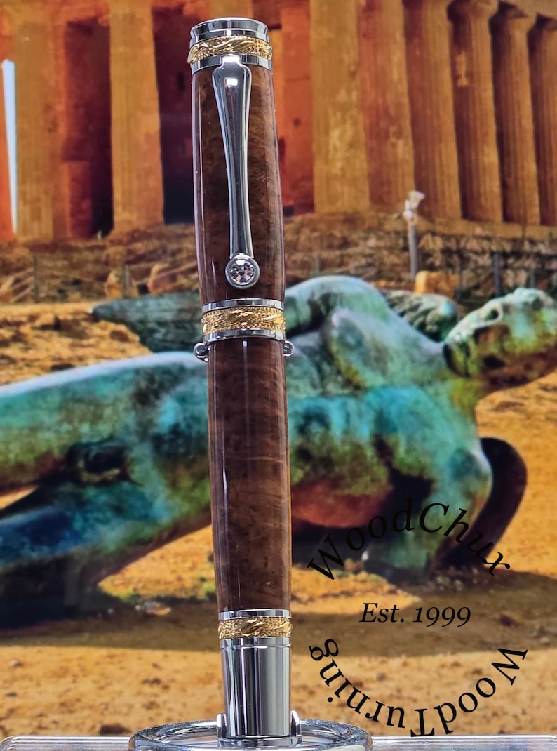 May include: A handcrafted wooden pen with a silver clip and gold accents. The pen features a dark brown, textured barrel and a sparkling gem. The pen is displayed against a blurred background with the text "Wood Turning Est. 1999".