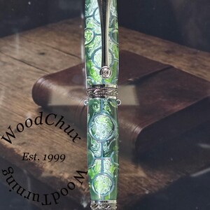 Może przedstawiać: Ręcznie wykonany długopis z zielono-białym wzorem na korpusie, srebrnymi akcentami i srebrnym klipsem. Długopis jest prezentowany na srebrnym stojaku. Długopis ma napis "WoodChux" i "Est. 1999" z boku.