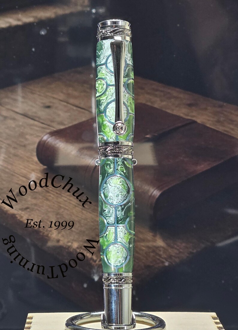 Może przedstawiać: Ręcznie wykonany długopis z zielono-białym wzorem na korpusie, srebrnymi akcentami i srebrnym klipsem. Długopis jest prezentowany na srebrnym stojaku. Długopis ma napis "WoodChux" i "Est. 1999" z boku.