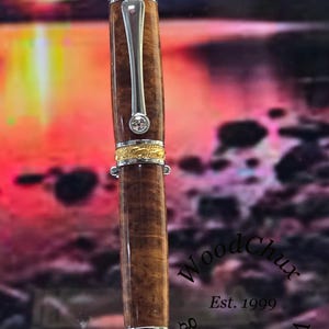 May include: A handcrafted wooden pen with a silver clip and accents. The pen features a brown, patterned barrel, gold-colored bands, and a small, clear gem. The pen is a WoodChux product, established in 1999.