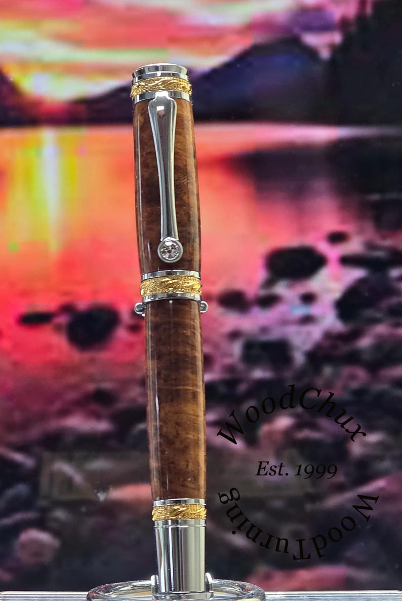 May include: A handcrafted wooden pen with a silver clip and accents. The pen features a brown, patterned barrel, gold-colored bands, and a small, clear gem. The pen is a WoodChux product, established in 1999.