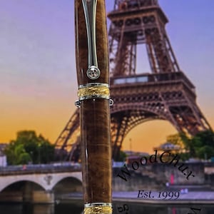 May include: A handcrafted wooden pen with a silver clip and gold accents. The pen features a brown, textured barrel and a sparkling gem. The background shows the Eiffel Tower and the text "WoodChux Est. 1999".