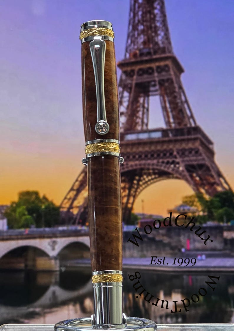 May include: A handcrafted wooden pen with a silver clip and gold accents. The pen features a brown, textured barrel and a sparkling gem. The background shows the Eiffel Tower and the text "WoodChux Est. 1999".