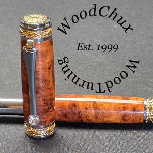 May include: A handcrafted pen with a wooden barrel featuring a unique burl grain pattern. The pen has silver and gold-colored metal accents, including a decorative cap and clip. The pen is lying on a dark surface.