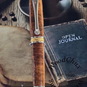 May include: A handcrafted wooden pen with a silver clip and accents. The pen features a brown, patterned barrel, gold-colored bands, and a small, clear gem. The pen is displayed with a book and coffee cup in the background.