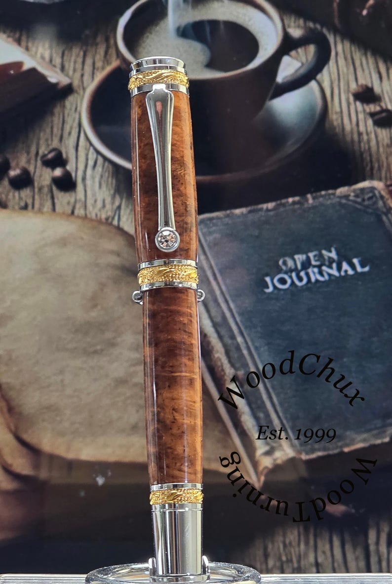 May include: A handcrafted wooden pen with a silver clip and accents. The pen features a brown, patterned barrel, gold-colored bands, and a small, clear gem. The pen is displayed with a book and coffee cup in the background.