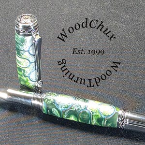 Może przedstawiać: Ręcznie wykonany długopis z zielono-białym marmurkowym wzorem, srebrnymi akcentami i ozdobną nasadką. Długopis jest pokazany bez nasadki, odsłaniając końc&oacute;wkę do pisania. Obraz zawiera tekst "WoodChux Est. 1999".