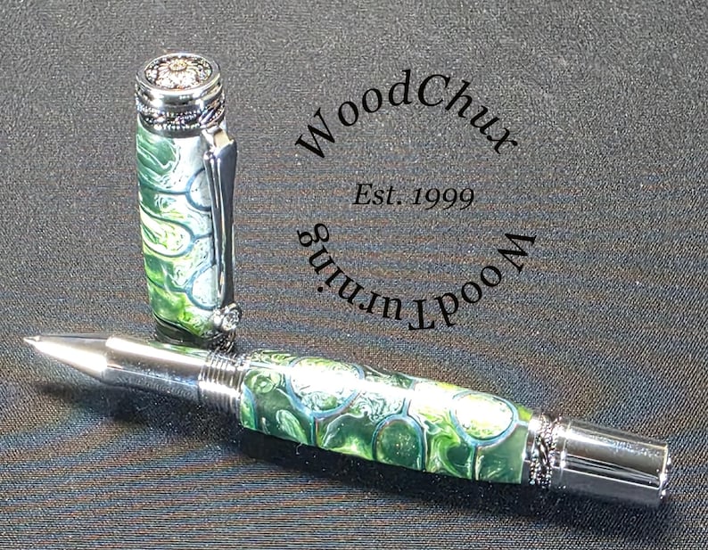 Może przedstawiać: Ręcznie wykonany długopis z zielono-białym marmurkowym wzorem, srebrnymi akcentami i ozdobną nasadką. Długopis jest pokazany bez nasadki, odsłaniając końc&oacute;wkę do pisania. Obraz zawiera tekst "WoodChux Est. 1999".