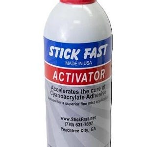 Puede incluir: Un bote de aerosol blanco con tapa roja, etiquetado como STICK FAST ACTIVATOR. La etiqueta presenta bandas azules y rojas, texto que indica que acelera el adhesivo de cianoacrilato, información de contacto y un peso neto de 354 g.