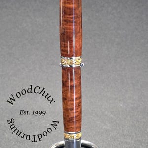 May include: A handcrafted wooden pen with a rich, brown burl wood grain. The pen features gold and silver accents and is displayed on a silver pen stand. The text "WoodChux Est. 1999" is visible.