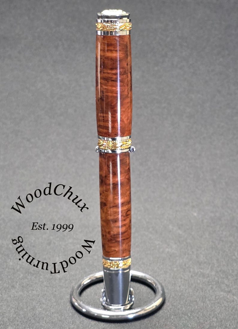 May include: A handcrafted wooden pen with a rich, brown burl wood grain. The pen features gold and silver accents and is displayed on a silver pen stand. The text "WoodChux Est. 1999" is visible.