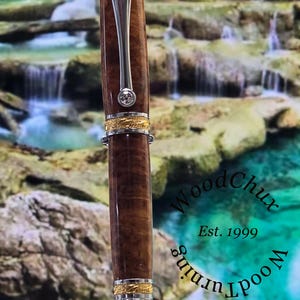 May include: A handcrafted wooden pen with a polished finish, featuring gold-colored accents and a silver clip. The pen has a clear, sparkling gem near the top. The text "WoodChux Est. 1999" is visible.