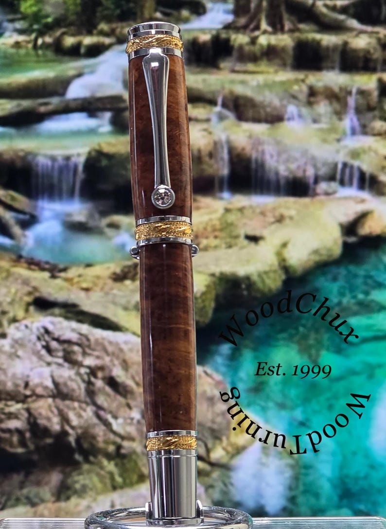 May include: A handcrafted wooden pen with a polished finish, featuring gold-colored accents and a silver clip. The pen has a clear, sparkling gem near the top. The text "WoodChux Est. 1999" is visible.