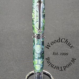 Może przedstawiać: Ręcznie wykonany długopis z zielono-białym wzorem na korpusie i srebrnymi akcentami. Długopis jest prezentowany na srebrnym stojaku na długopisy. Widoczny jest tekst "WoodChux Woodturning Est. 1999".