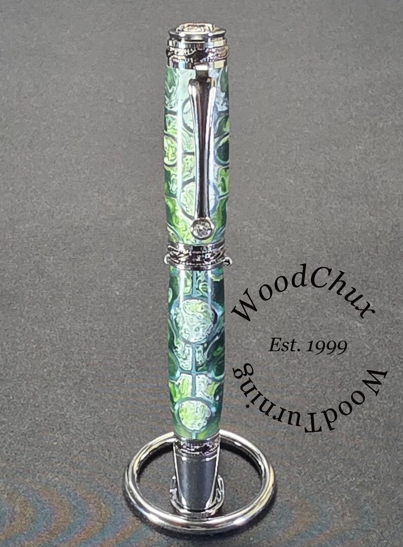Może przedstawiać: Ręcznie wykonany długopis z zielono-białym wzorem na korpusie i srebrnymi akcentami. Długopis jest prezentowany na srebrnym stojaku na długopisy. Widoczny jest tekst "WoodChux Woodturning Est. 1999".
