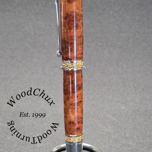 May include: A handcrafted wooden pen with a polished, reddish-brown finish and gold-colored accents. The pen features a silver clip and is displayed on a silver pen stand. The pen's body has a unique wood grain pattern. The text "WoodChux" and "Est. 1999" are visible.