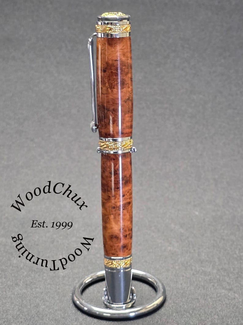 May include: A handcrafted wooden pen with a polished, reddish-brown finish and gold-colored accents. The pen features a silver clip and is displayed on a silver pen stand. The pen's body has a unique wood grain pattern. The text "WoodChux" and "Est. 1999" are visible.
