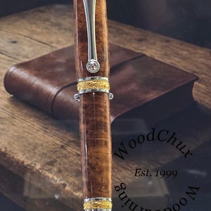 May include: A handcrafted wooden pen with a polished finish, accented with gold-colored bands and a silver clip. The pen features a clear gemstone and is displayed with a leather notebook in the background. The pen is approximately 6 inches long.