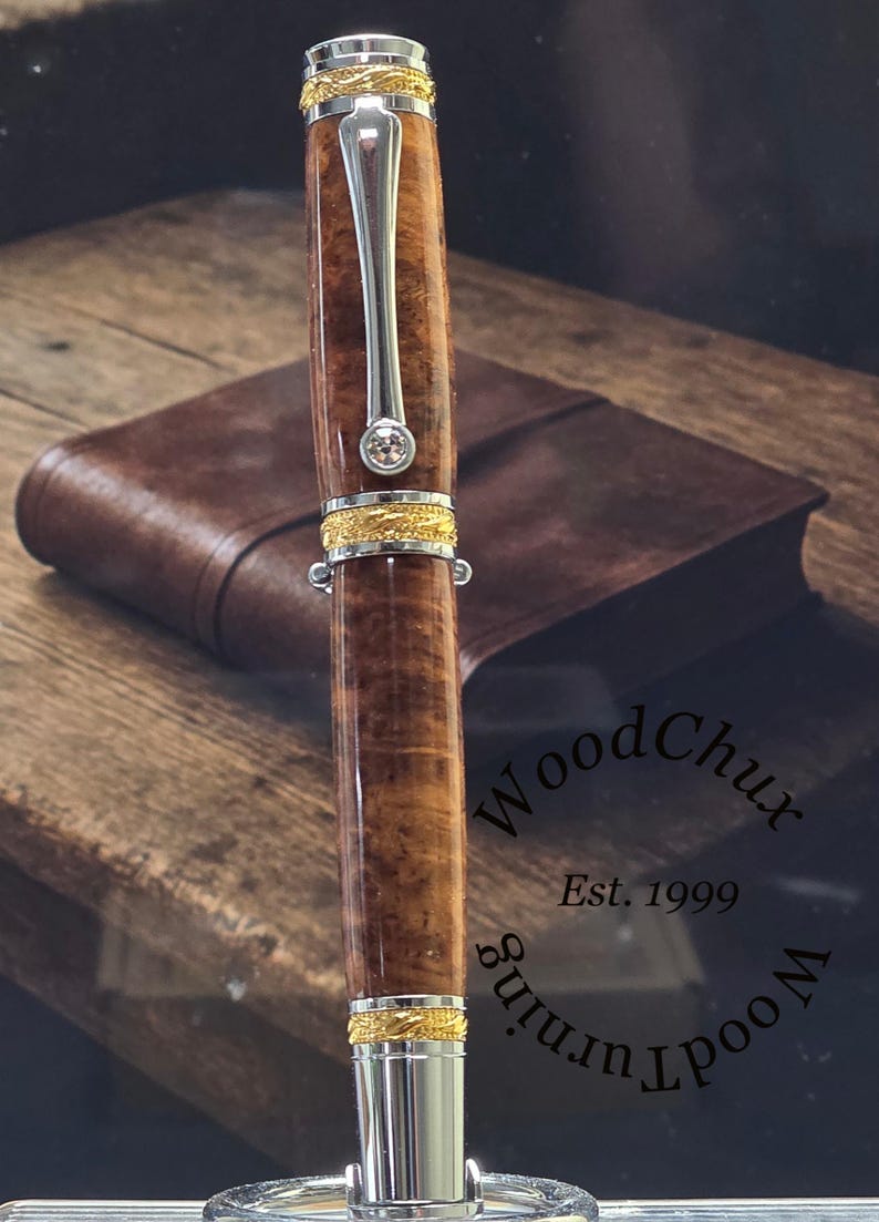 May include: A handcrafted wooden pen with a polished finish, accented with gold-colored bands and a silver clip. The pen features a clear gemstone and is displayed with a leather notebook in the background. The pen is approximately 6 inches long.
