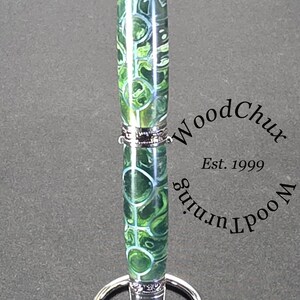 Może przedstawiać: Ręcznie wykonany długopis z zielono-białym wzorem w wiry, zaakcentowany srebrnym metalem. Długopis jest prezentowany na srebrnym stojaku na długopisy. Długopis ma napis "WoodChux Wood Turning Est. 1999".