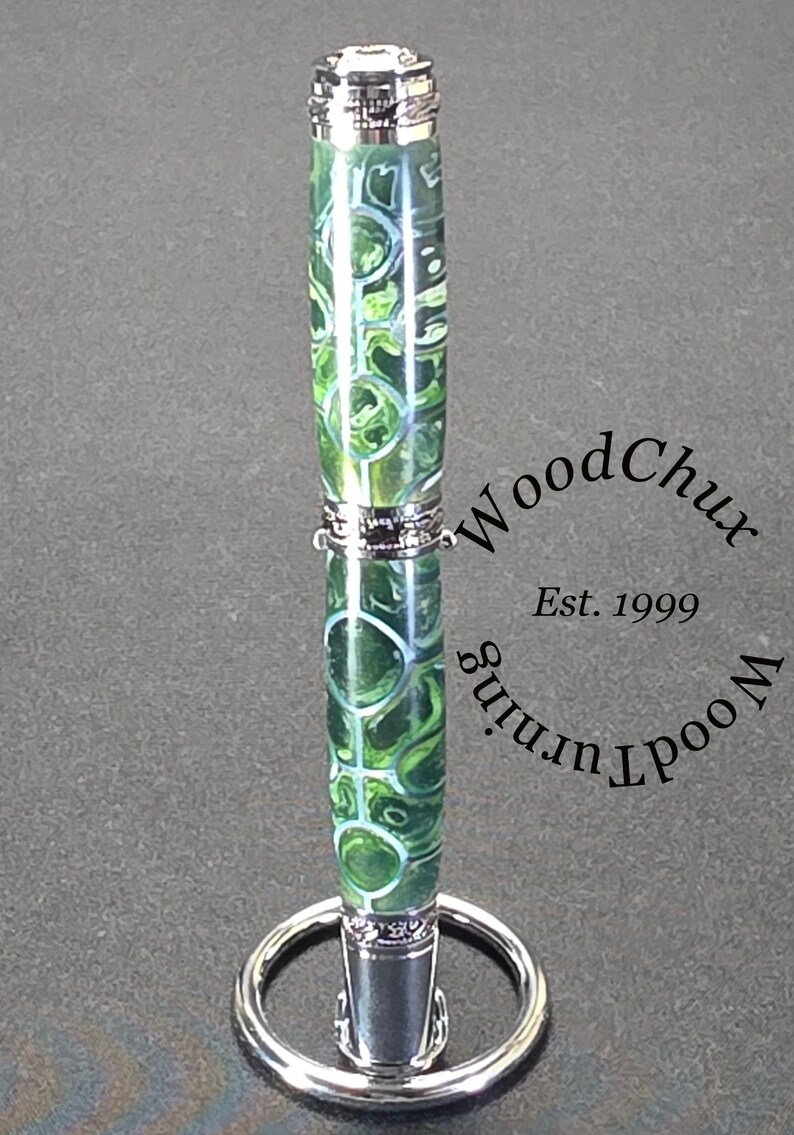 Może przedstawiać: Ręcznie wykonany długopis z zielono-białym wzorem w wiry, zaakcentowany srebrnym metalem. Długopis jest prezentowany na srebrnym stojaku na długopisy. Długopis ma napis "WoodChux Wood Turning Est. 1999".