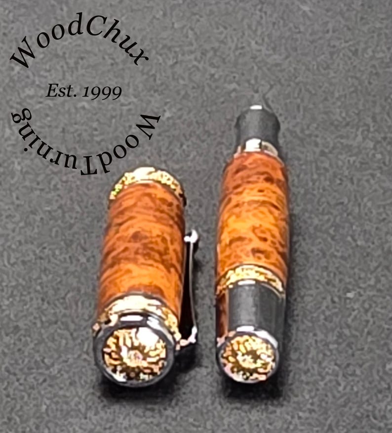 May include: A handcrafted fountain pen with a wooden barrel and a silver-colored clip. The pen's barrel has a unique wood grain pattern with gold accents. The pen cap is detached, revealing the nib. The text "WoodChux" and "Est. 1999" are visible.