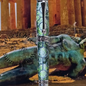 Może przedstawiać: Ręcznie wykonany długopis z zielono-białym marmurkowym wzorem, zaakcentowany srebrnym metalem. Długopis posiada klips i ozdobne paski. Długopis jest prezentowany na stojaku. Widoczny jest tekst "WoodChux" i "Est. 1999".