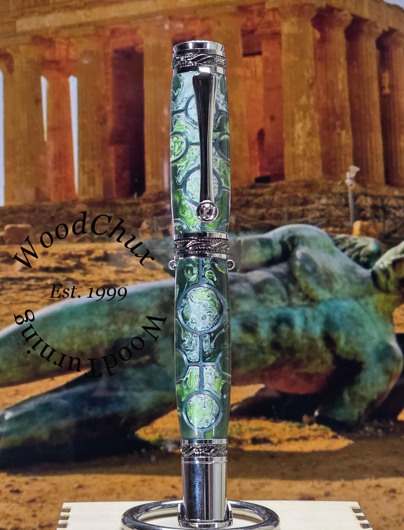 Może przedstawiać: Ręcznie wykonany długopis z zielono-białym marmurkowym wzorem, zaakcentowany srebrnym metalem. Długopis posiada klips i ozdobne paski. Długopis jest prezentowany na stojaku. Widoczny jest tekst "WoodChux" i "Est. 1999".