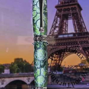 Może przedstawiać: Ręcznie wykonany długopis z zielono-białym wzorem na korpusie, srebrnymi akcentami i ozdobnym klipsem. Długopis jest prezentowany na rozmytym tle z Wieżą Eiffla i tekstem "WoodChux Est. 1999".