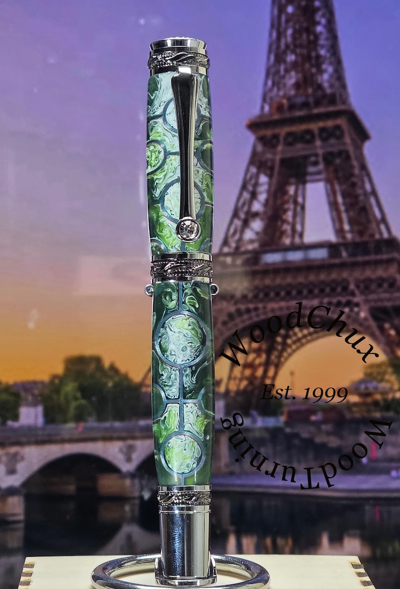 Może przedstawiać: Ręcznie wykonany długopis z zielono-białym wzorem na korpusie, srebrnymi akcentami i ozdobnym klipsem. Długopis jest prezentowany na rozmytym tle z Wieżą Eiffla i tekstem "WoodChux Est. 1999".