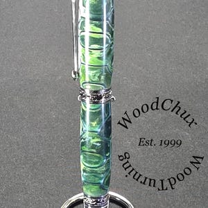 Może przedstawiać: Ręcznie wykonany długopis z żywym zielono-niebieskim wzorem na korpusie, zaakcentowany srebrnymi metalowymi elementami. Długopis jest prezentowany na srebrnym stojaku. Widoczny jest tekst "WoodChux, Est. 1999".