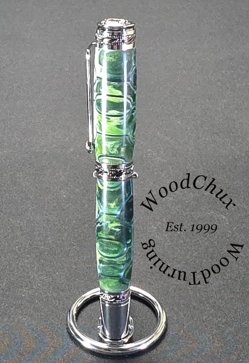 Może przedstawiać: Ręcznie wykonany długopis z żywym zielono-niebieskim wzorem na korpusie, zaakcentowany srebrnymi metalowymi elementami. Długopis jest prezentowany na srebrnym stojaku. Widoczny jest tekst "WoodChux, Est. 1999".