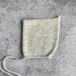 May include: A light gray knitted baby bonnet with long ties. The bonnet is made of a soft, textured yarn and has a square crown. The ties are long and can be tied under the chin. The bonnet is shown on a gray background.