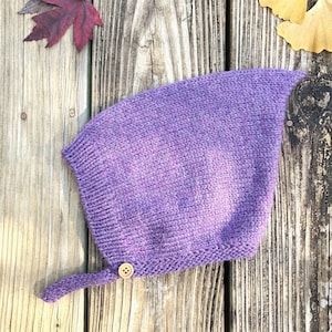 May include: A knitted purple bonnet with a button closure and a small tail. The bonnet is made of a soft, textured yarn. The background is a weathered wooden surface with autumn leaves.