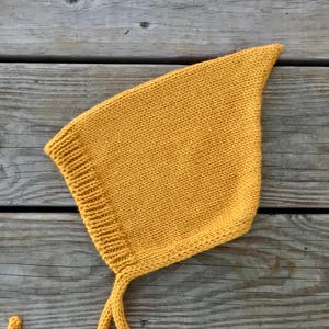 May include: A mustard yellow knitted baby bonnet with a long tie. The bonnet is lying on a wooden surface.