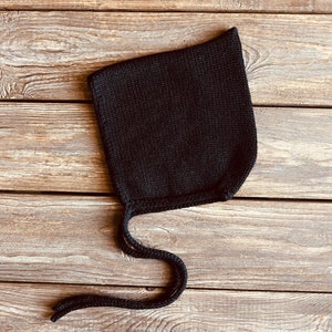 May include: A black knitted baby bonnet with a long tie. The bonnet is lying on a wooden surface.