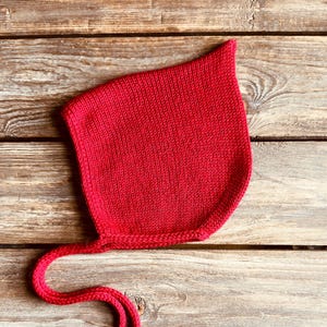 May include: A bright red knitted baby bonnet with long ties. The bonnet is made of a soft, textured yarn and is designed to cover the head and neck. The ties are used to secure the bonnet under the chin. The bonnet is displayed on a wooden surface.