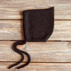 May include: A black knitted baby bonnet with a long tie. The bonnet is lying on a wooden surface.