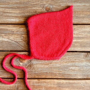 May include: A bright red knitted baby bonnet with long ties. The bonnet is made of a soft, textured yarn and is designed to cover the head and neck. The ties are used to secure the bonnet under the chin.