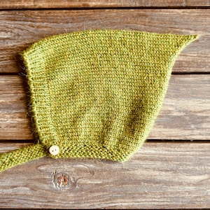 May include: A knitted, olive green bonnet with a pointed top and a button closure. The bonnet has a long, knitted tie on one side. The item is displayed on a rustic, wooden surface.