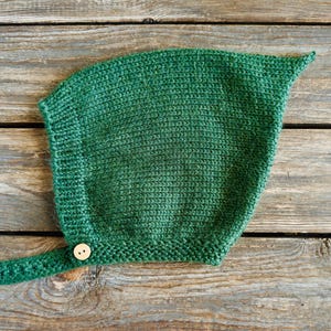 May include: A forest green knitted bonnet featuring a pointed top and a ribbed border along the face opening. The bonnet includes a single chin strap secured by a small, round, light-colored wooden button with two visible holes in the center.
