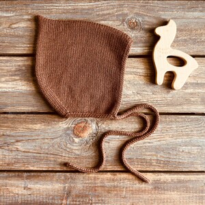 May include: A brown knitted baby bonnet with tie strings. The bonnet is made of a soft, textured yarn. A wooden deer-shaped toy is next to the bonnet. The items are on a wooden surface.