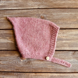 May include: A knitted baby bonnet in a dusty rose color. The bonnet features a pointed top, a button closure, and a strap. The item is made of a soft, textured yarn and is designed to keep a baby's head warm.