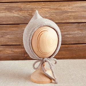 May include: A light gray knitted baby bonnet with a pointed top and a braided edge. The bonnet is tied with a matching ribbon. The bonnet is displayed on a wooden head form against a wooden background.