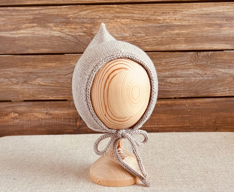 May include: A light gray knitted baby bonnet with a pointed top and a braided edge. The bonnet is tied with a matching ribbon. The bonnet is displayed on a wooden head form against a wooden background.