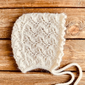 May include: A white crocheted baby bonnet with a delicate lace pattern and a long tie. The bonnet is lying on a wooden surface.